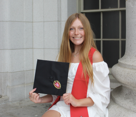 Ava Heyrman wearing a white dress and red graduation stole, smiling while holding a black graduation cap with a University of Wisconsin emblem.