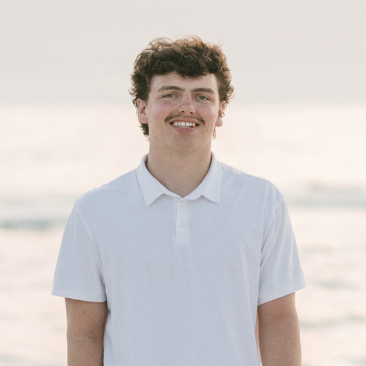 Headshot of CJ Varney smiling outdoors in a white polo shirt with a beach in the background.