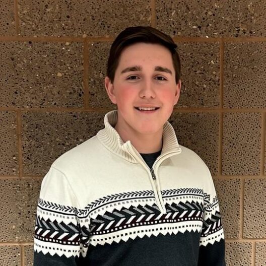 Headshot of Isaac Schultz smiling in a patterned half-zip sweater against a brown brick wall.