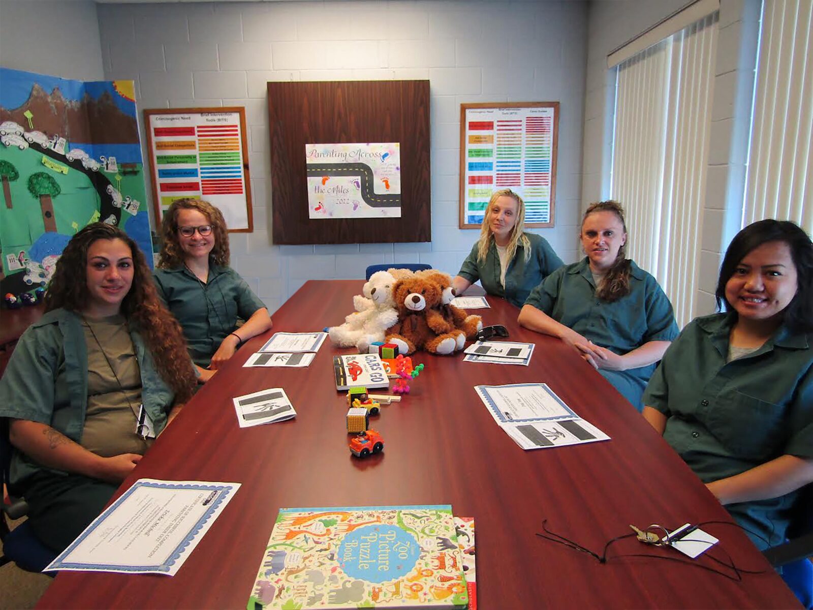 Five participants sitting around a conference table filled with children's books, toys, and stuffed animals under a poster that reads Parenting Across the Miles.