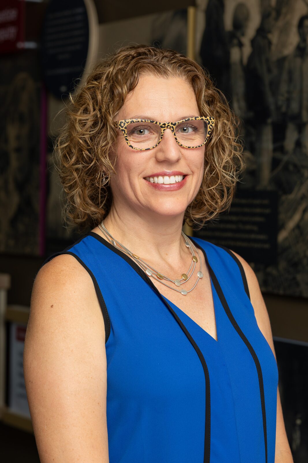 Portrait of Pajarita Charles smiling, wearing leopard-print glasses and a blue sleeveless shirt with black trim.