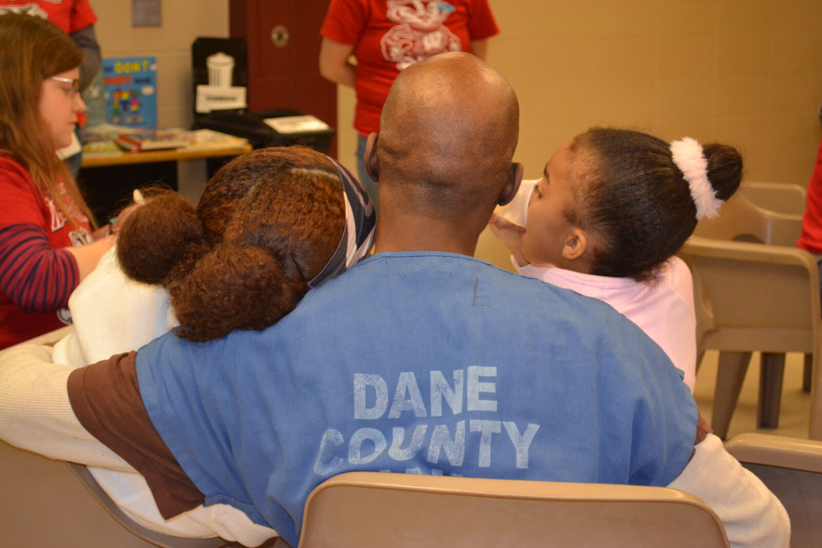 A parent in a blue Dane County Jail shirt hugging two young children seated together from behind during a visitation.